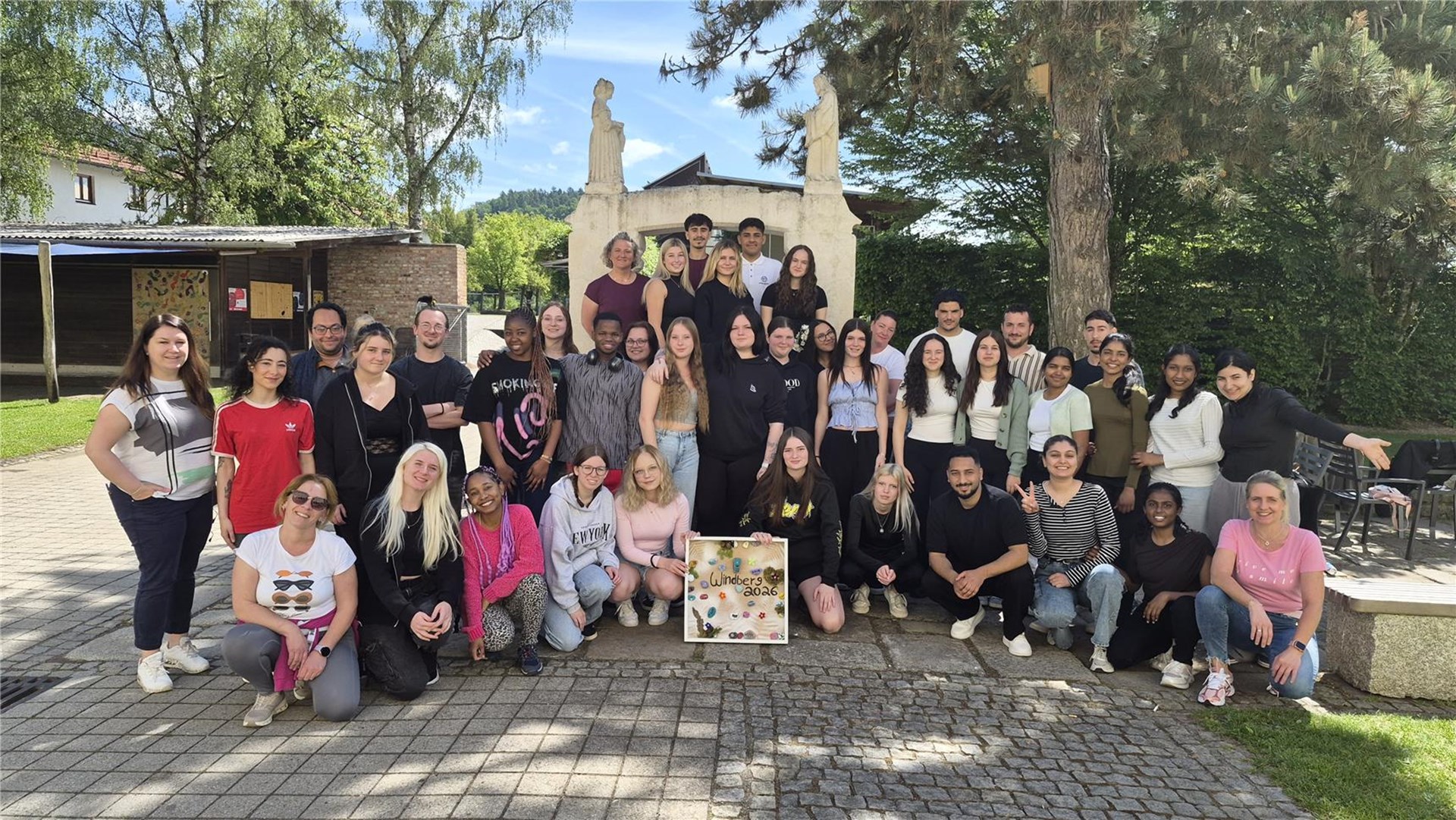 Eine große Gruppe von Schülerinnen und Schülern sowie Mitarbeitenden der Berufsfachschule für Pflege in Windberg steht gemeinsam vor einem Gebäude im Grünen. In der Mitte hält die Gruppe ein dekoriertes Schild mit der Aufschrift „Windenberg 2026“.